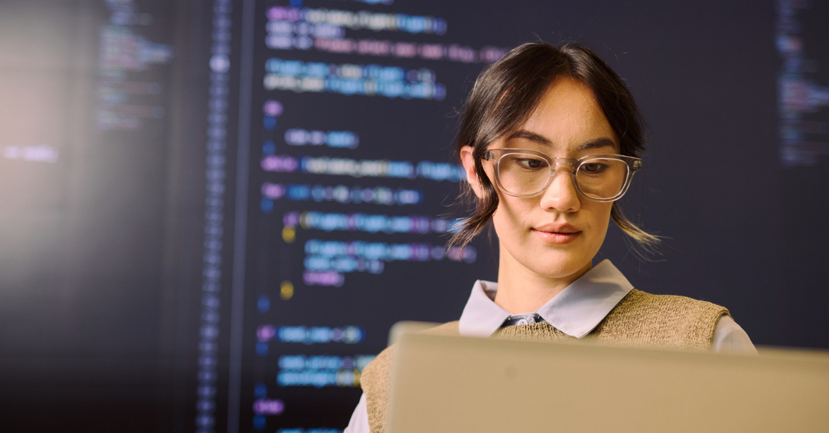A young professional wearing glasses works on a laptop with colorful code displayed on a large screen in the background.